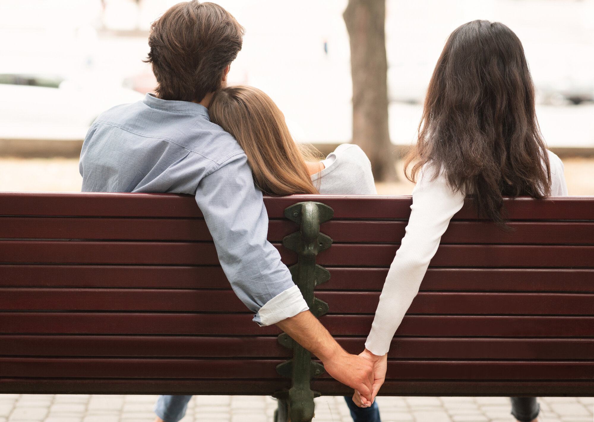 Husband Holding Hands With Girlfriend's Friend Sitting On Bench Outdoor